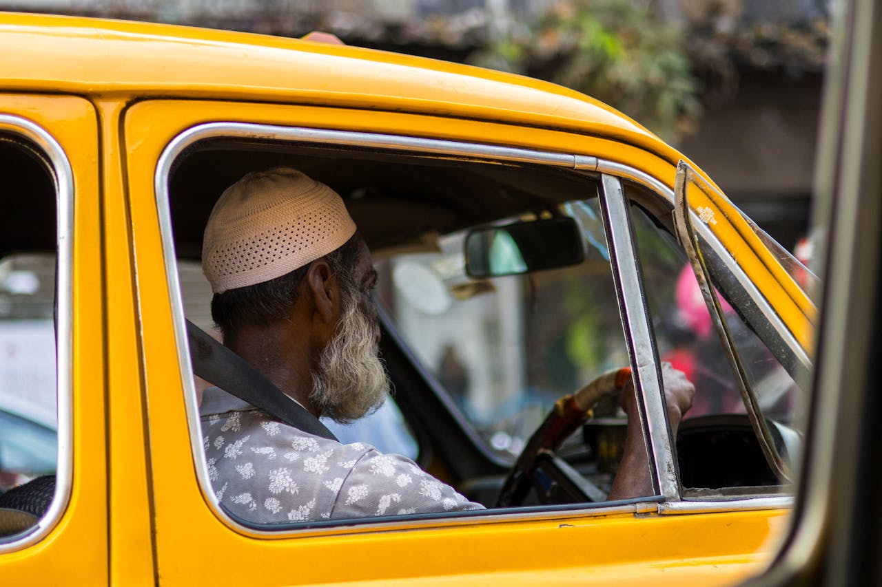 services-01 Elderly man in a yellow taxi driving through an urban street, wearing a traditional cap.