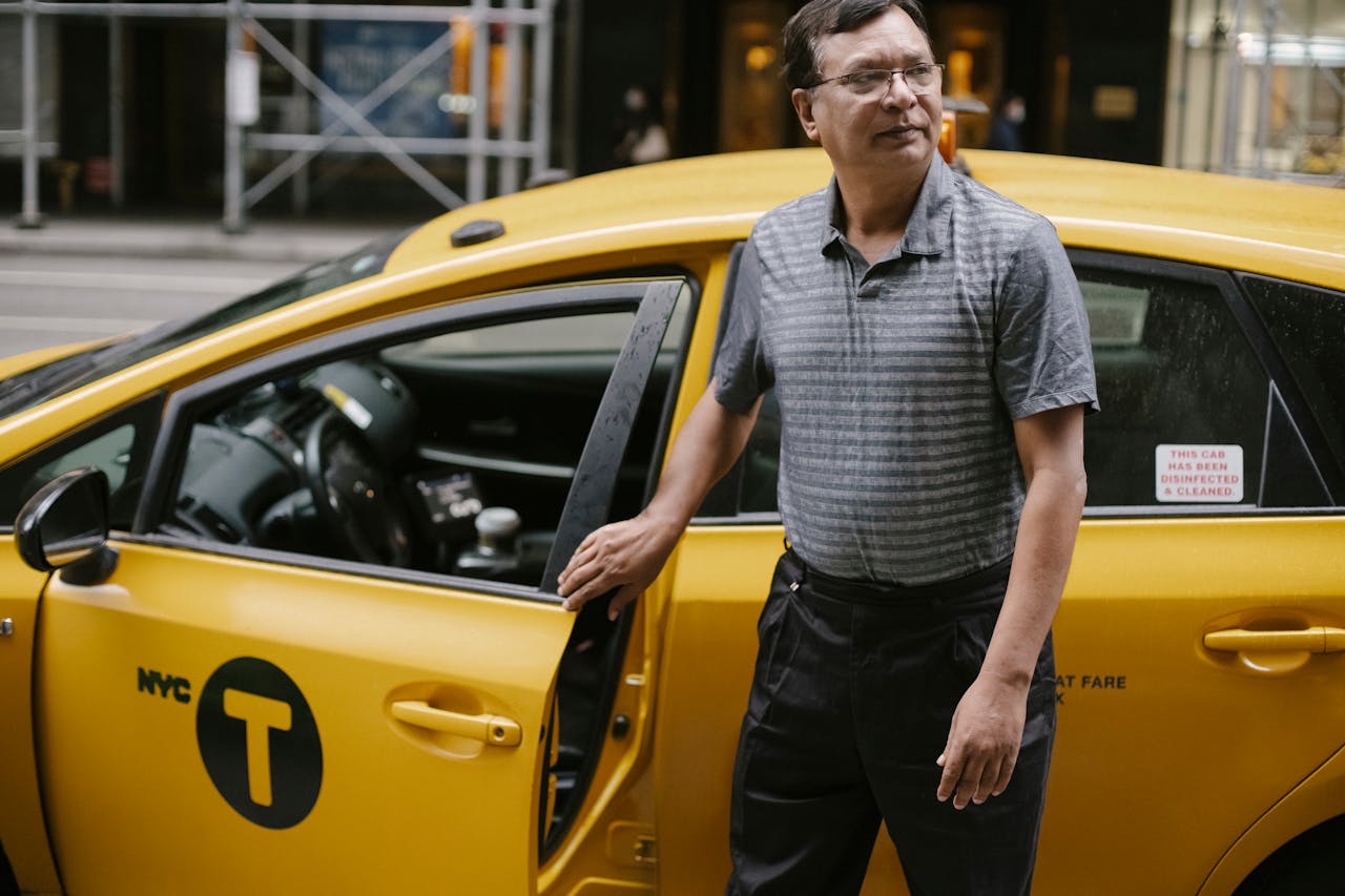 services-04 Serious crop ethnic male in casual clothes and eyeglasses standing near modern yellow taxi parked on street in New York and looking away